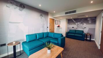 A modern living room with teal sofas, a wooden coffee table, and a large wall mural of a cricket player.