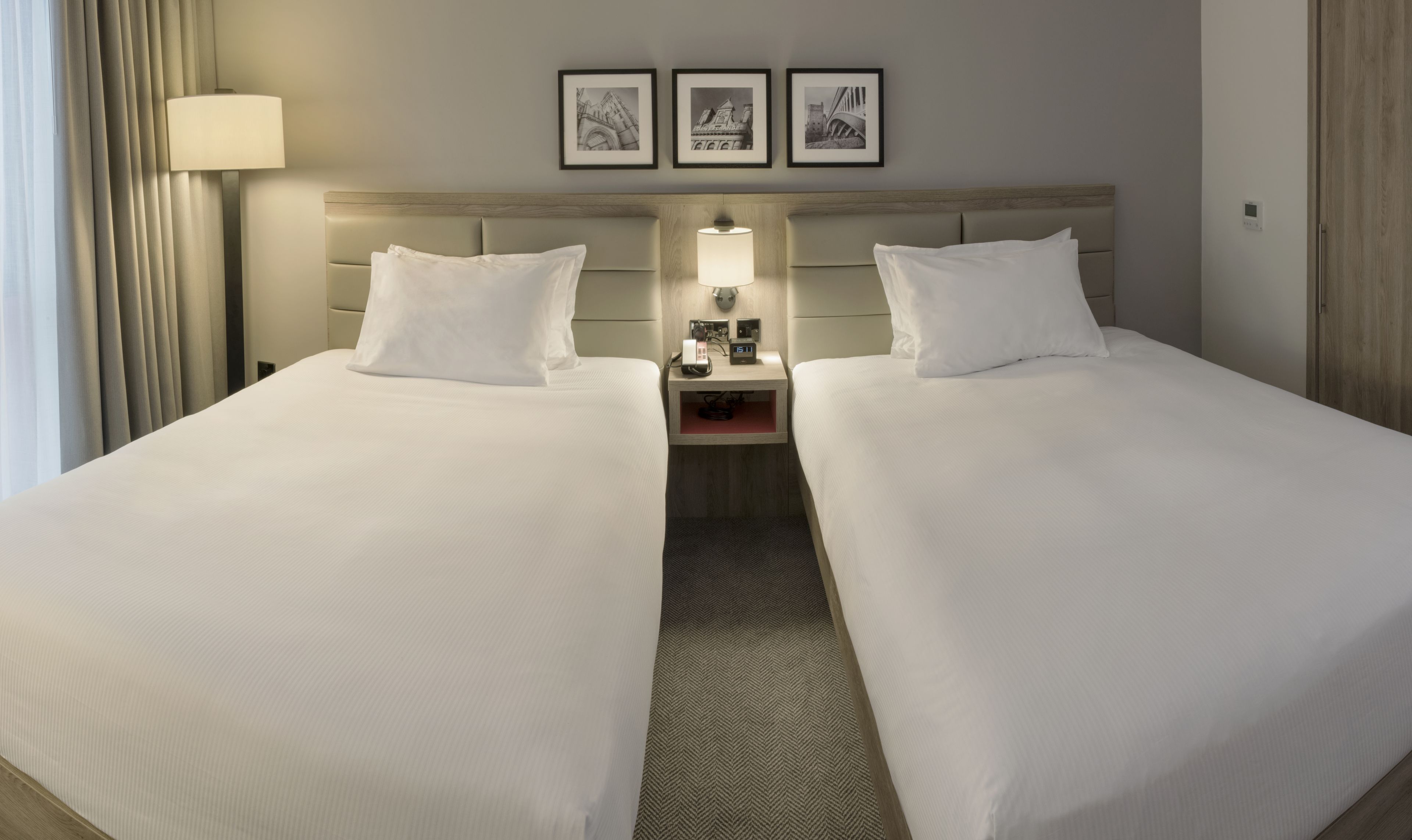 Een hotelkamer met twee bedden, een nachtkastje met een lamp, een klok en drie ingelijste foto's aan de muur.