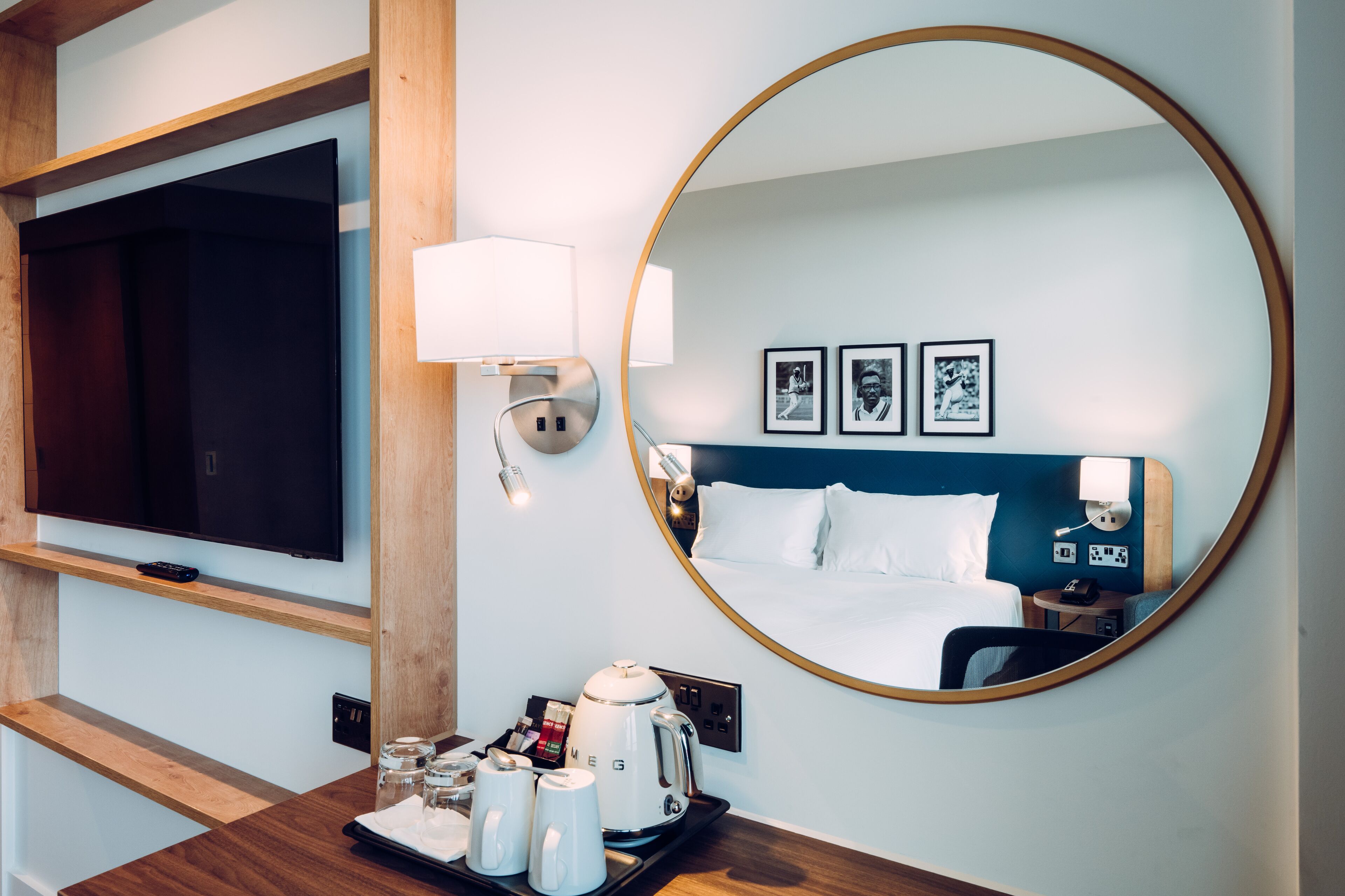 A hotel room with a large mirror, a flat-screen TV, a bed with white linens, a bedside table with a lamp, and a tray with coffee-making items.