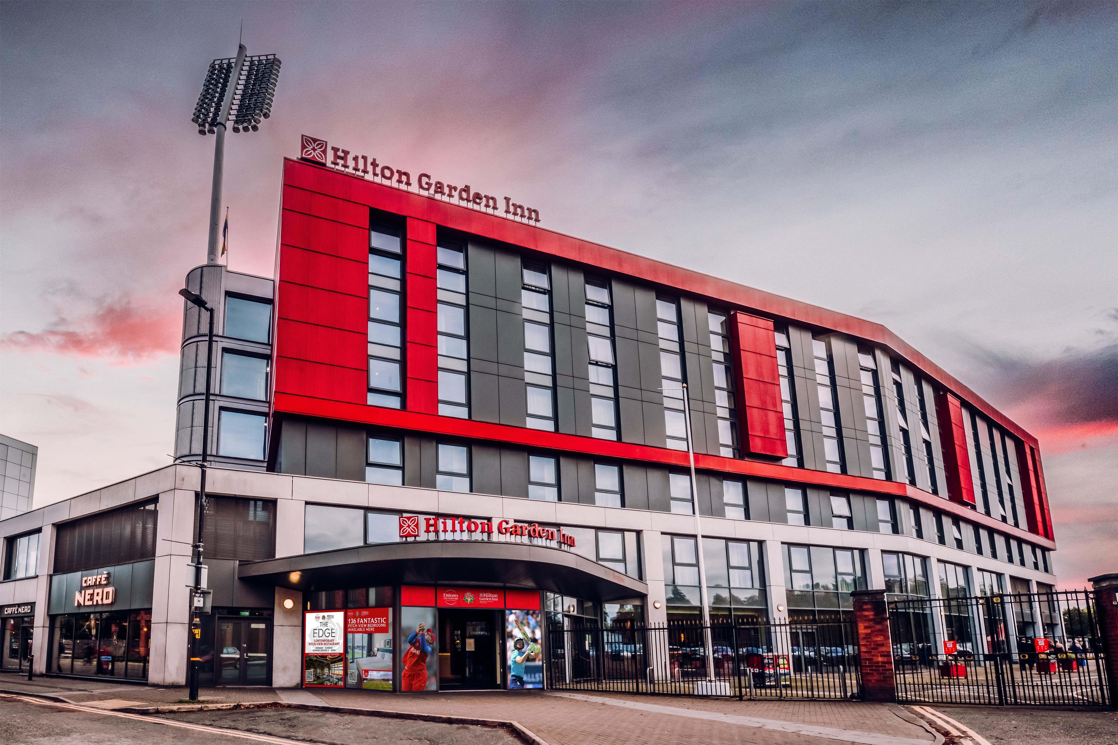 A modern Hilton Garden Inn hotel with a prominent red and grey facade, large windows, and a visible entrance with signage.