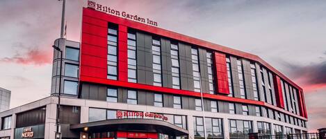 A modern Hilton Garden Inn hotel with a prominent red and grey facade, large windows, and a visible entrance with signage.