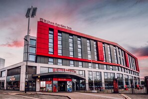 A modern Hilton Garden Inn hotel with a prominent red and grey facade, large windows, and a visible entrance with signage.