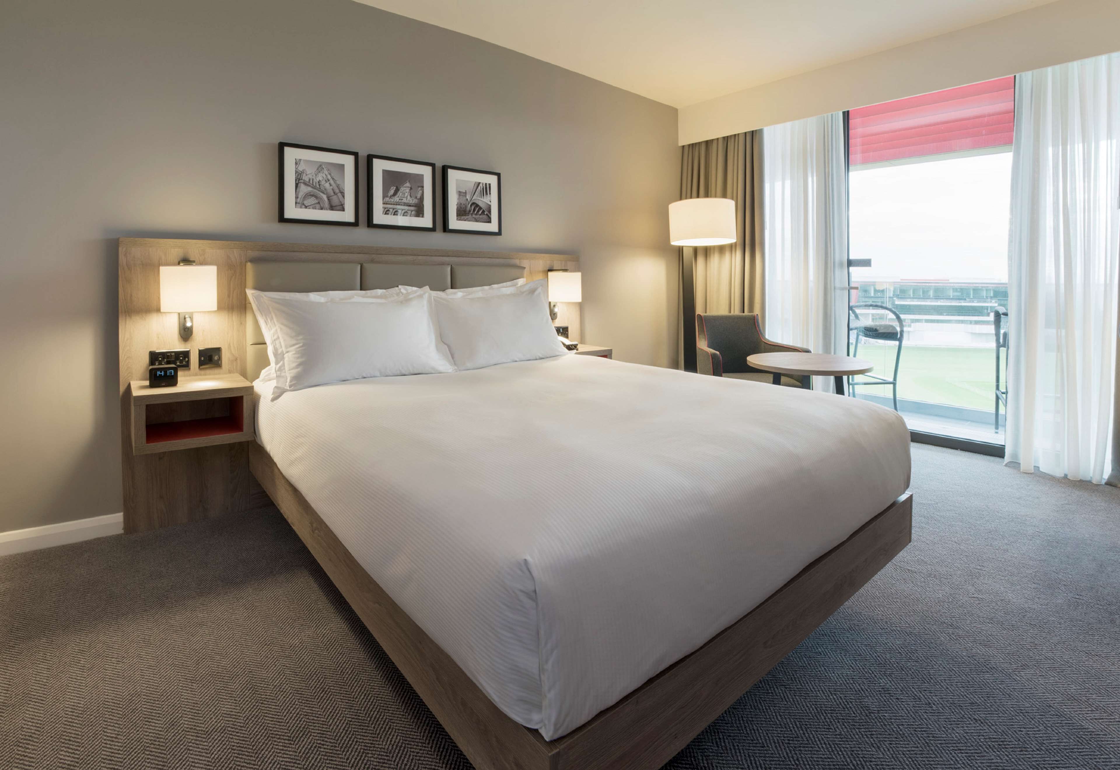 A hotel room with a large bed, two bedside tables with lamps, a small table and chair, and a balcony with a view.