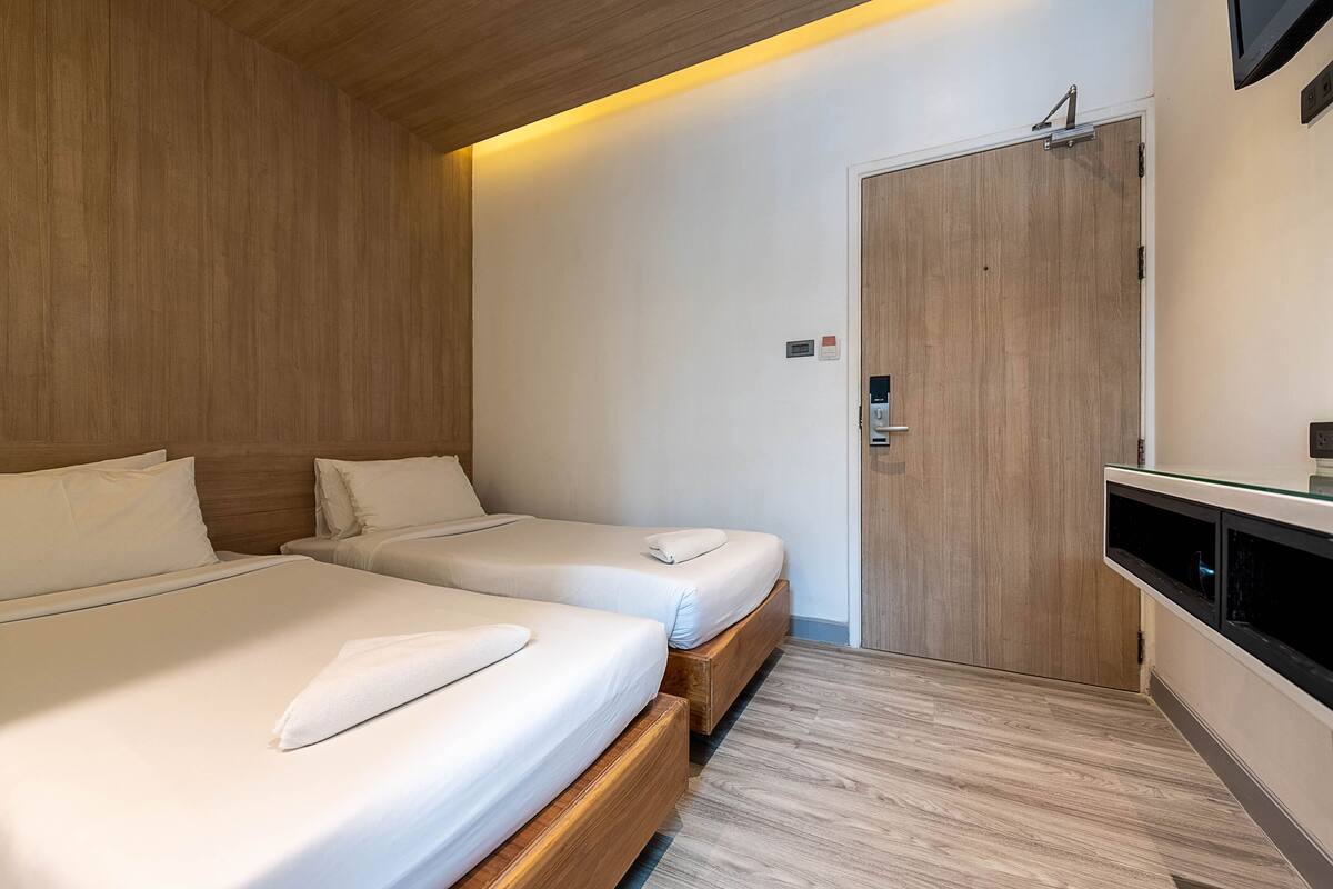 A hotel room with two beds, wooden flooring, a built-in bench, a TV, and a door with a lock.