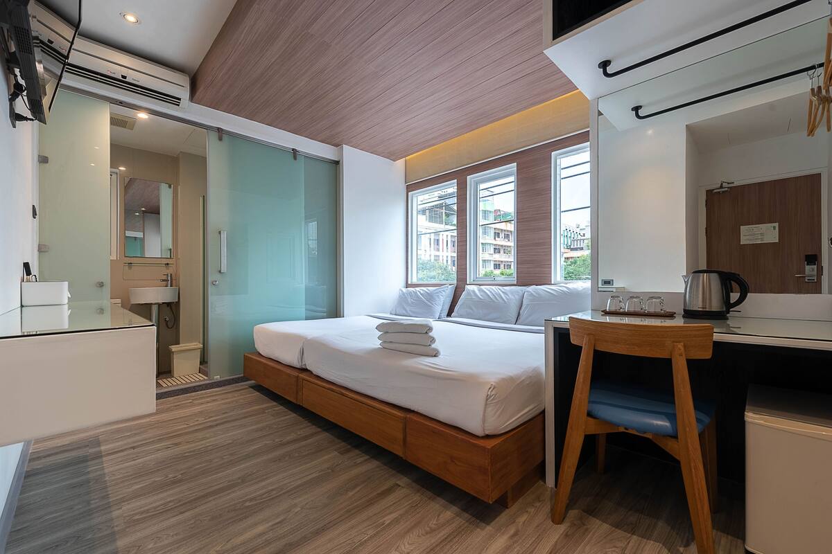 A modern hotel room with a bed, a desk, and a view of buildings through large windows.