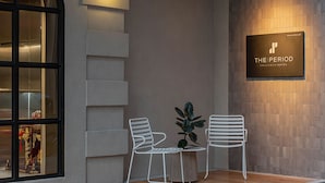 A modern hotel entrance with a sign reading "THE PERIOD PRATUNAM HOTEL". There are two white chairs and a small table with a plant. - The Period Pratunam (Bangkok)