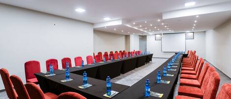 Un salón de conferencias con una mesa larga, sillas rojas y botellas de agua azules.