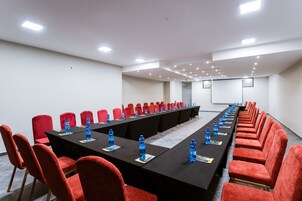 Uma sala de conferências com uma mesa comprida, cadeiras vermelhas e garrafas de água azuis.