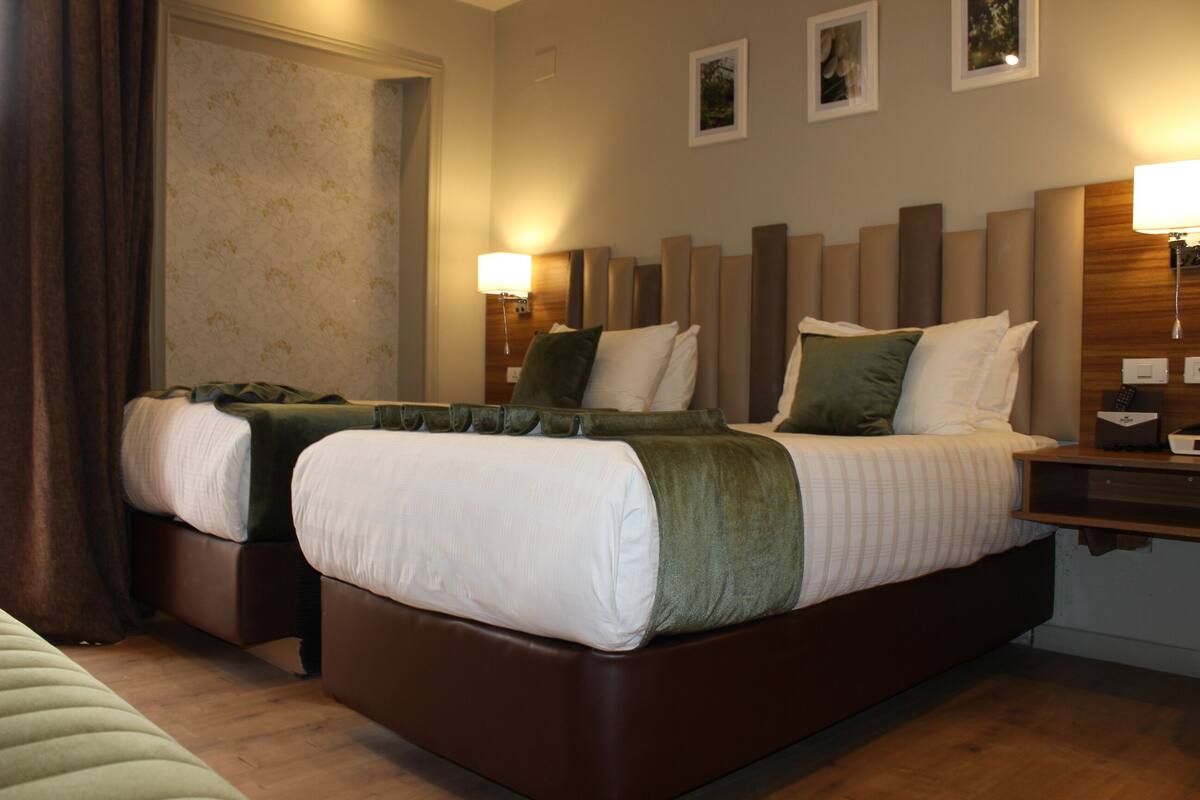 A hotel room with two beds, a wooden headboard, a nightstand, and a wall-mounted lamp.