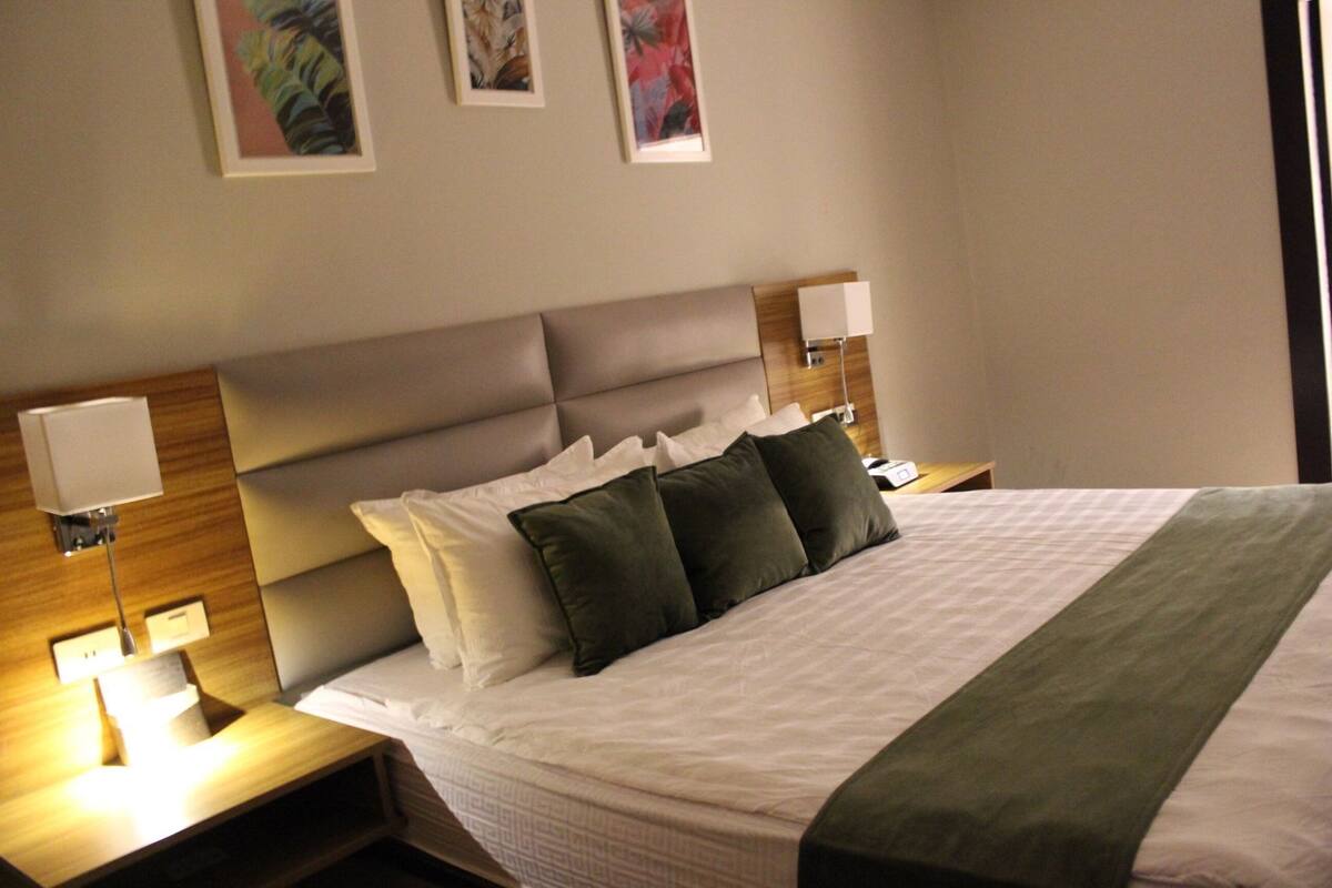 A hotel room with a large bed, two bedside tables with lamps, a headboard, and three framed artworks on the wall.