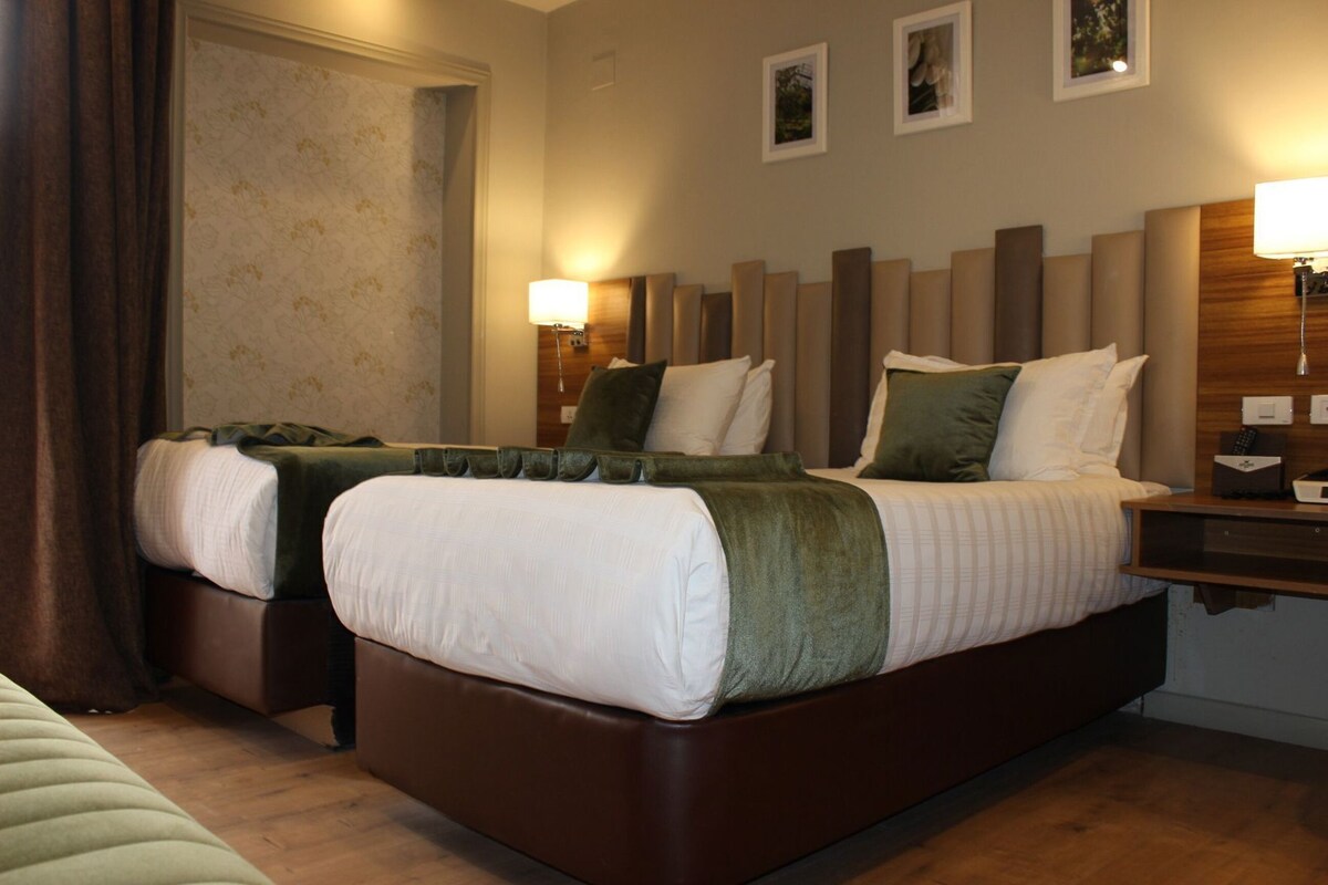 A hotel room with two beds, a wooden headboard, a bedside table with a lamp, and a wall-mounted light fixture.