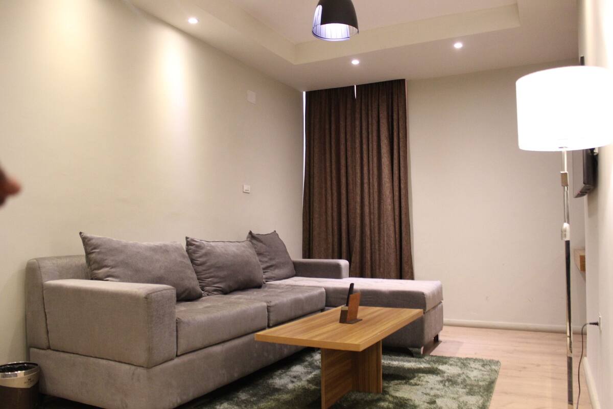 A modern living room with a grey sofa, a wooden coffee table, and a large window with curtains.