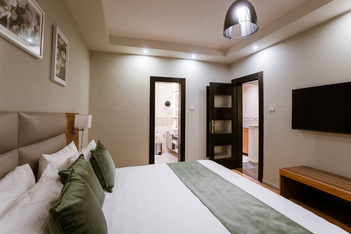 A modern hotel room with a large bed, a bedside table, a TV, and a door leading to a bathroom.