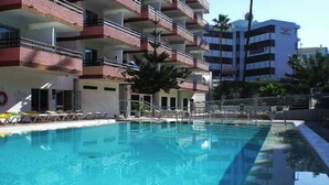 A swimming pool with a clear blue water surface, surrounded by a building with multiple balconies and a few palm trees. - Las Gacelas (San Bartolomé de Tirajana)