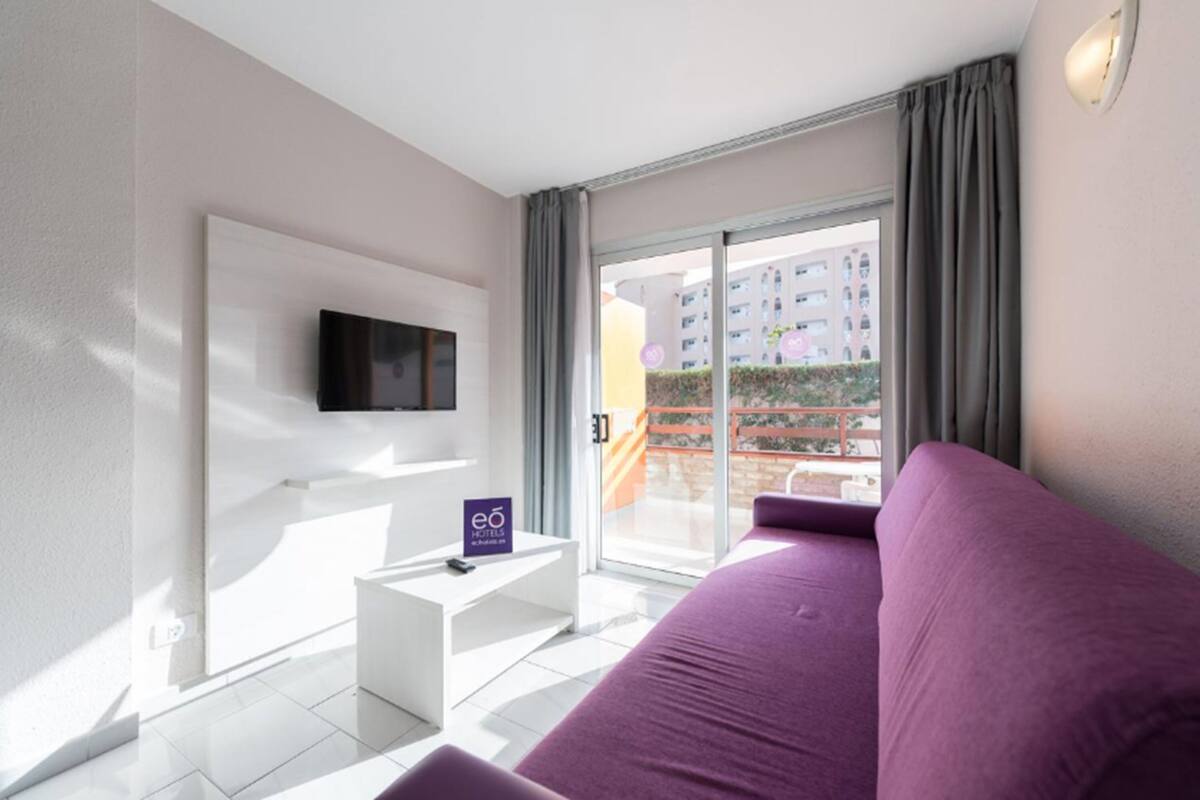 Une chambre d’hôtel moderne dotée d’une grande fenêtre, d’un téléviseur à écran plat fixé au mur, d’une petite table de chevet blanche et d’un canapé violet.
