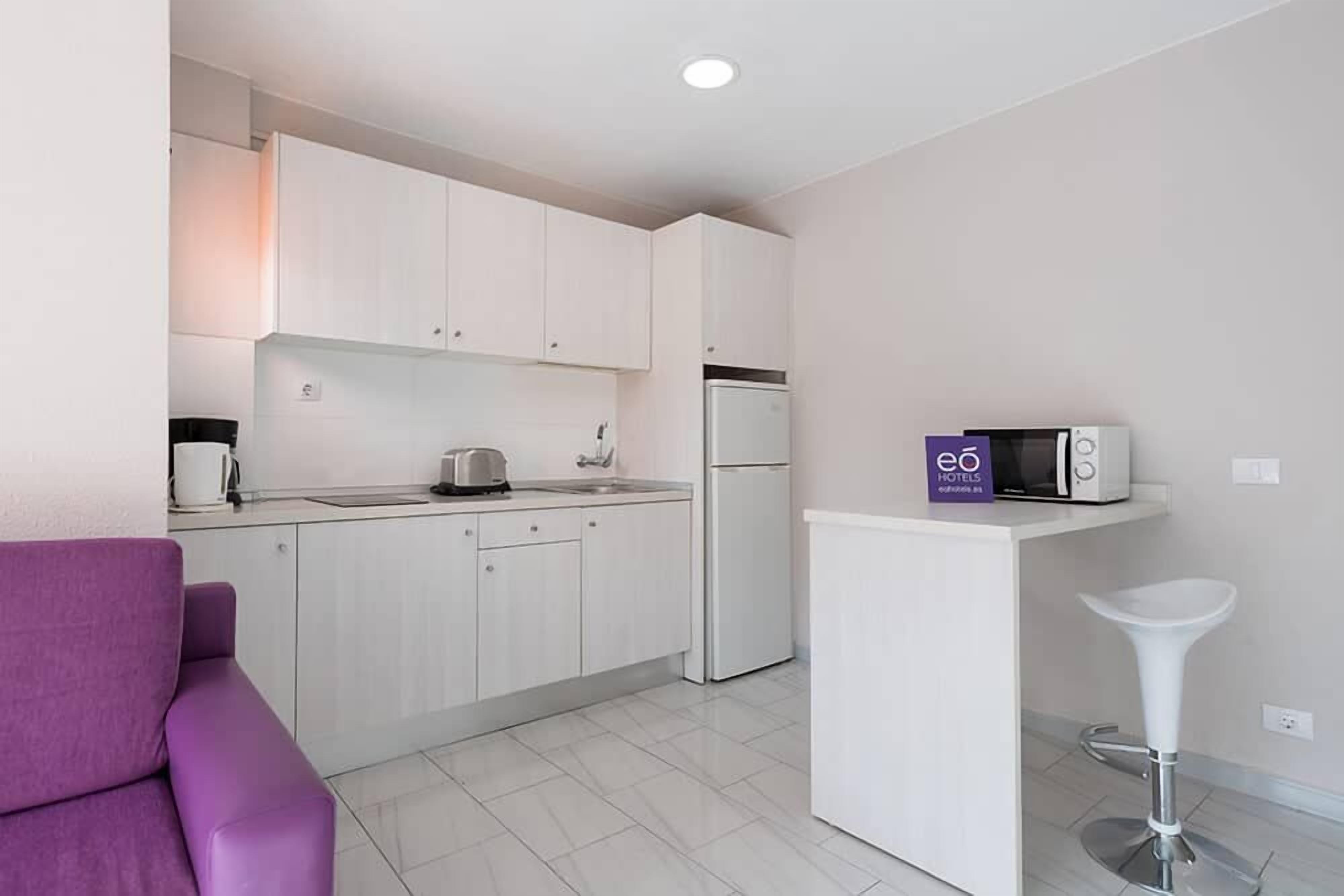 A modern kitchen with white cabinets, a refrigerator, a microwave, and a coffee maker. There is a purple sofa and a white bar stool.