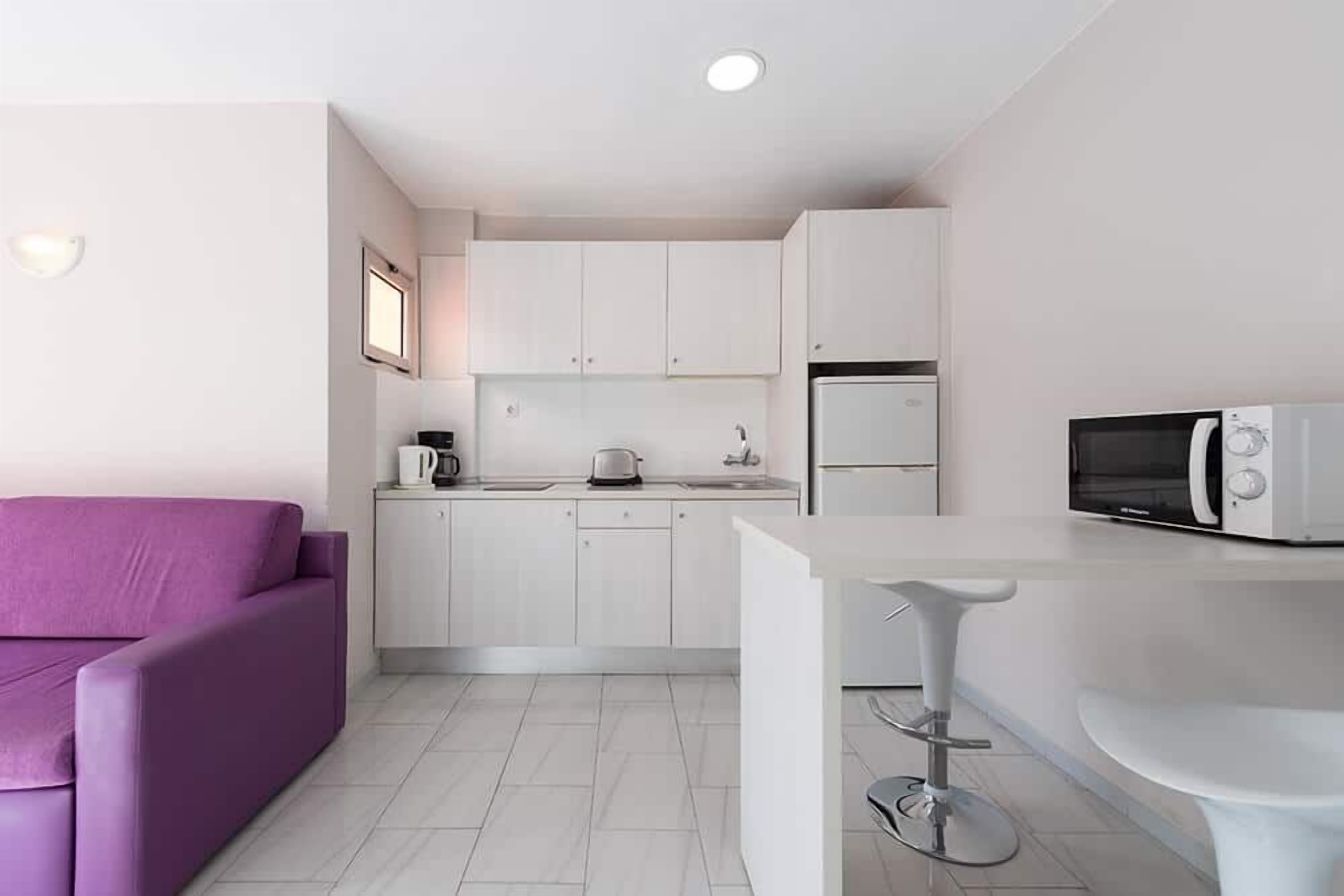 A compact kitchen with white cabinets, a microwave, a kettle, and a toaster. There is a purple sofa and a white bar stool.
