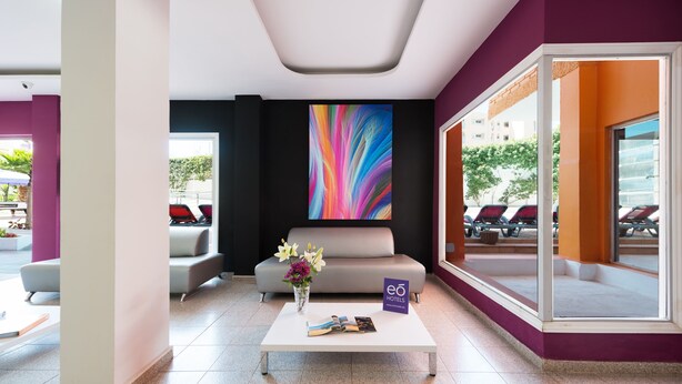 A hotel lobby with a white coffee table, a sofa, a colorful abstract painting, and a view of outdoor seating. - Las Gacelas (San Bartolomé de Tirajana)
