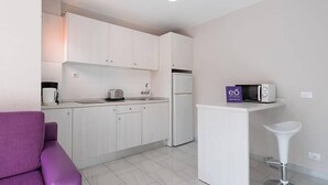 A modern kitchen with white cabinets, a refrigerator, a microwave, and a coffee maker. There is a purple sofa and a white bar stool. - Las Gacelas (San Bartolomé de Tirajana)