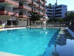 A swimming pool with a clear blue water surface, surrounded by a building with multiple balconies and a few palm trees.