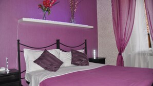 A bedroom with a bed, purple bedding, a nightstand, a lamp, and a shelf with flowers. - Belhorizon Guest House (Rome)
