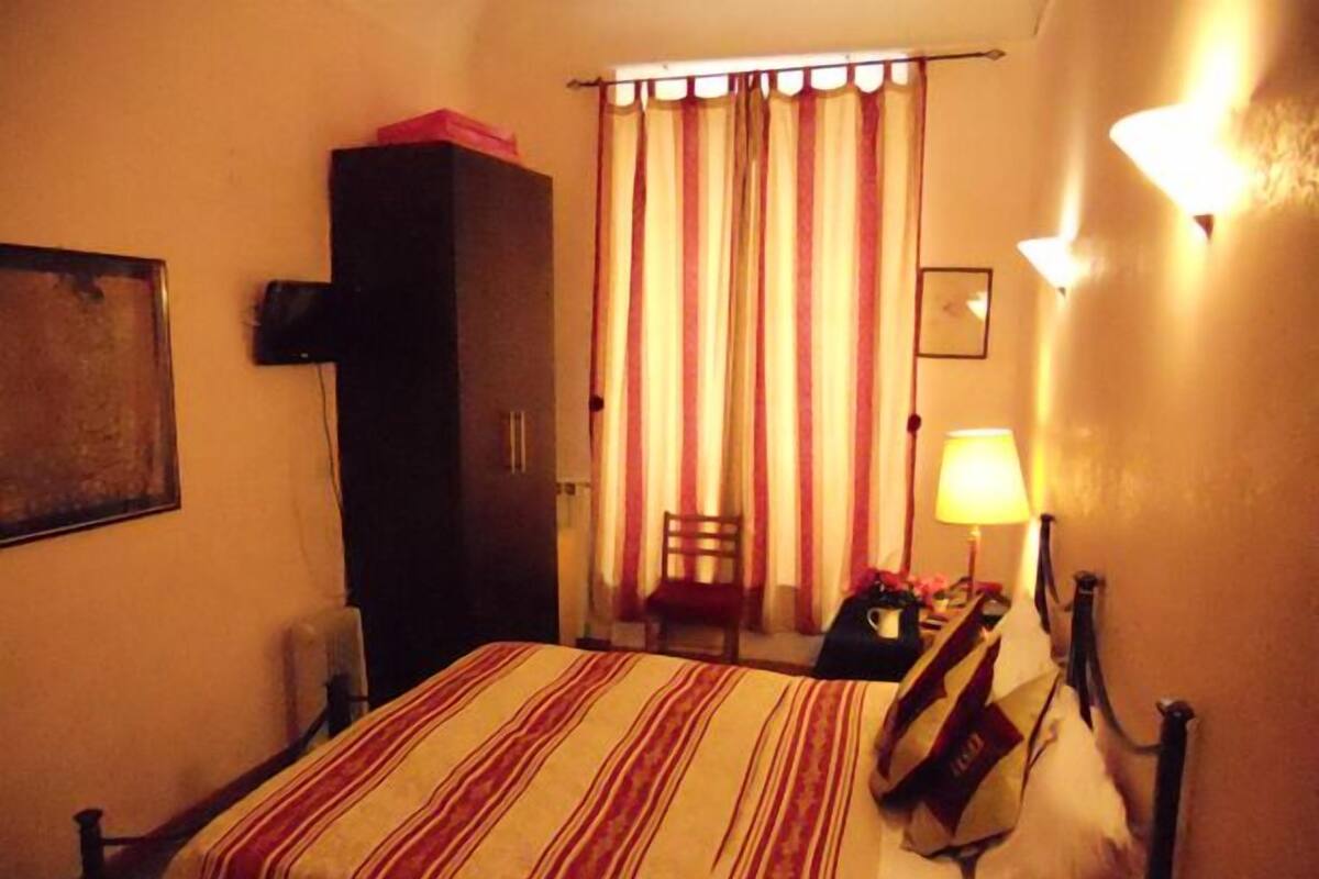 A hotel room with a bed, a chair, a desk with a lamp, and a window with curtains.