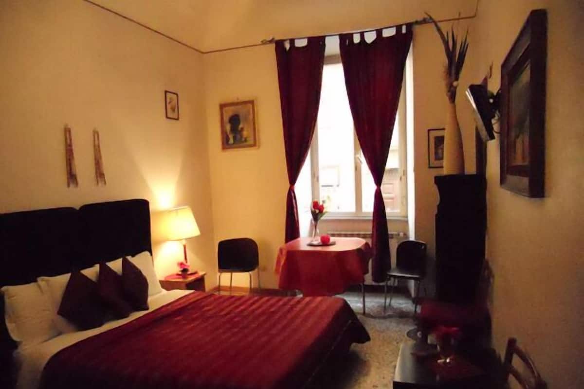 A hotel room with a bed, a desk, a chair, a table with a vase, and a window with curtains.