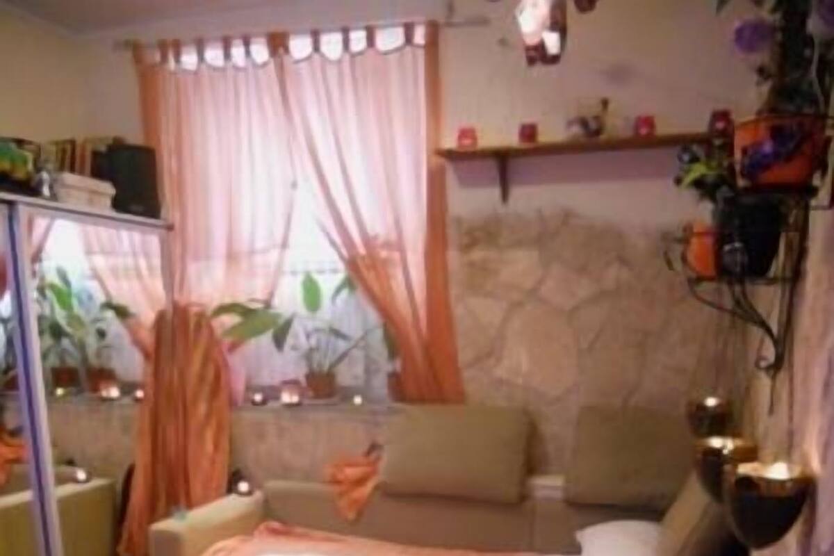 A bedroom with a bed, a sofa, a mirror, a small table with a candle, and a wall-mounted shelf with decorative items.