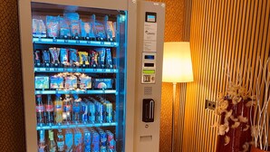 A vending machine stocked with various beverages and snacks, located in a room with a patterned wall and a lamp. - EuroStar Hotel (Castrop-Rauxel)