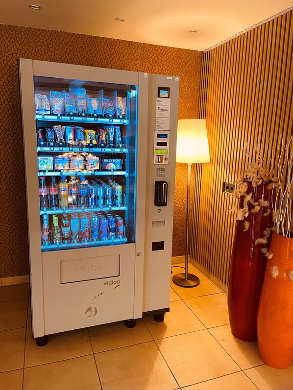 A vending machine stocked with various beverages and snacks, located in a room with a patterned wall and a lamp.