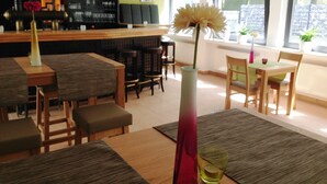 A modern bar area with a wooden counter and shelves of bottles. There are several tables and chairs arranged for dining. A tall vase with a flower is placed on a table in the foreground. - EuroStar Hotel (Castrop-Rauxel)