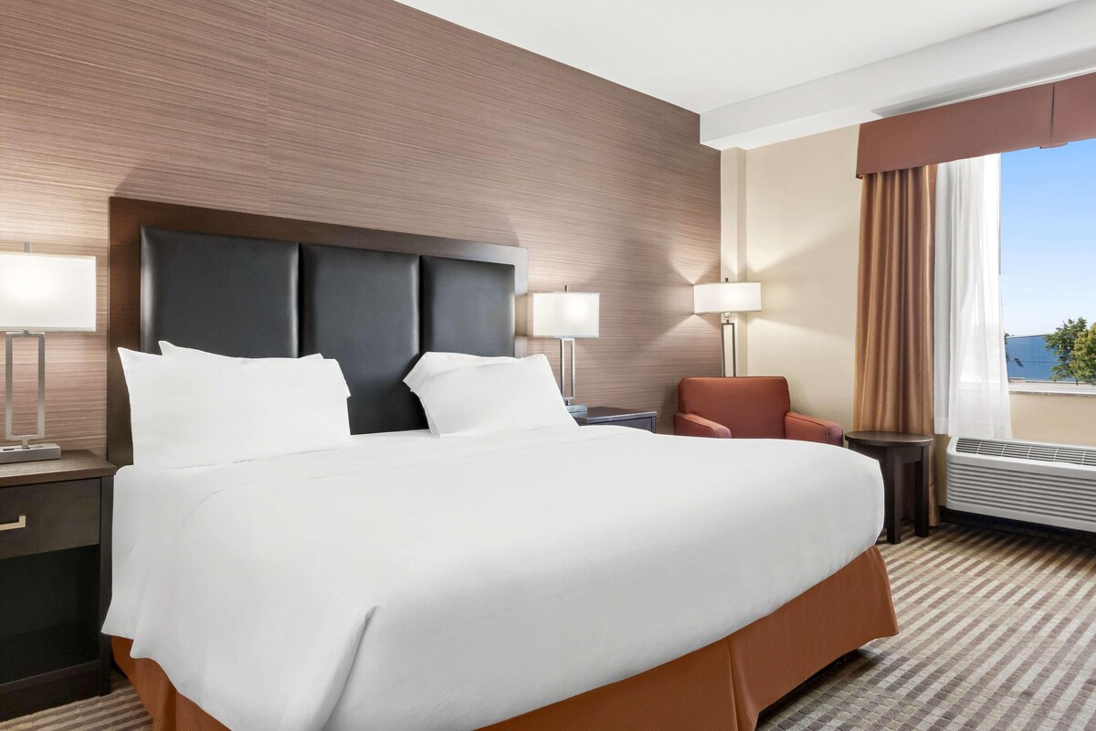 Habitación de hotel con una cama grande, una mesita de noche, una silla, una lámpara y vistas al exterior.