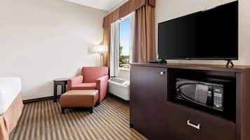A hotel room with a bed, a pink armchair, a television, a microwave, and a window with curtains.
