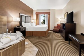 Habitación de hotel con cama, escritorio, televisor, lámpara de escritorio, silla, sofá, baño con bañera y ventana con cortinas.