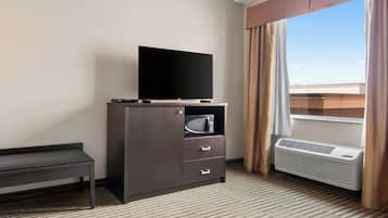 A hotel room with a flat-screen TV on a cabinet, a microwave, a bench, and a large window with curtains.