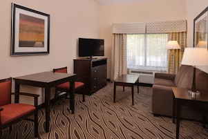 Habitación de hotel con una mesa de comedor, dos sillas, un sofá, un televisor y una ventana con cortinas.