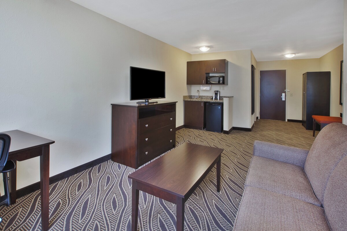 Een hotelkamer met een flatscreen televisie, een kitchenette met een magnetron, een koffiezetapparaat en een kleine koelkast. Er is een bank en een tafel met een kamerbreed tapijt.