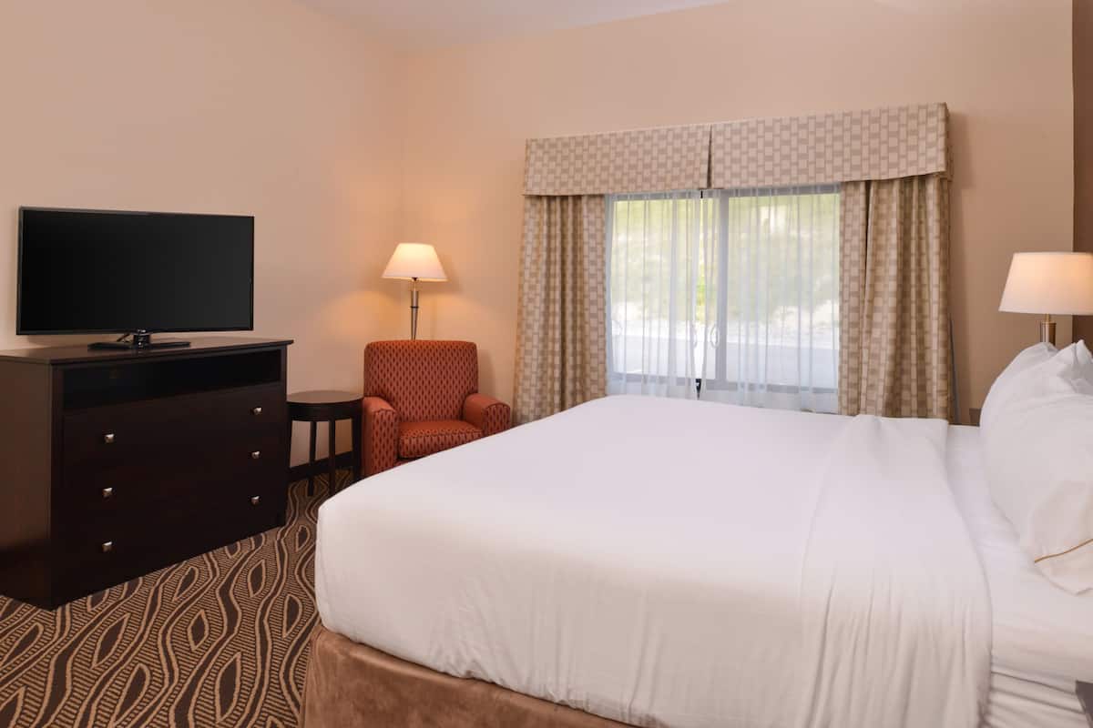 A hotel room with a large bed, a TV on a dresser, a chair, and a window with curtains.
