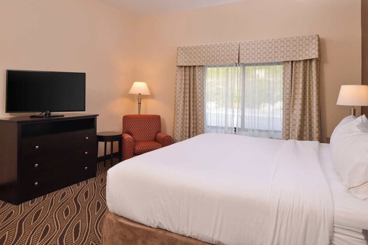Een hotelkamer met een groot bed, een flatscreen televisie op een dressoir, een stoel en een raam met gordijnen.