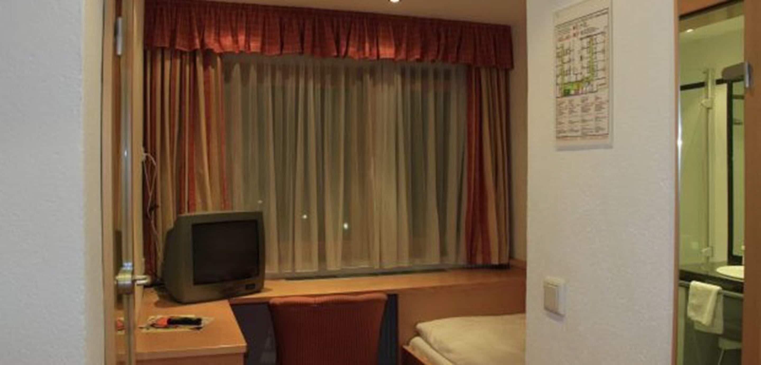 Una camera d'albergo con un letto, una televisione, una scrivania con una sedia e una finestra con tende.