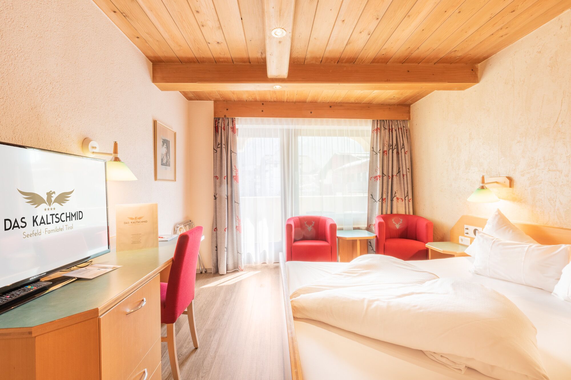 A hotel room with a wooden ceiling, a bed, a desk with a television, a red chair, and a small table with a lamp.