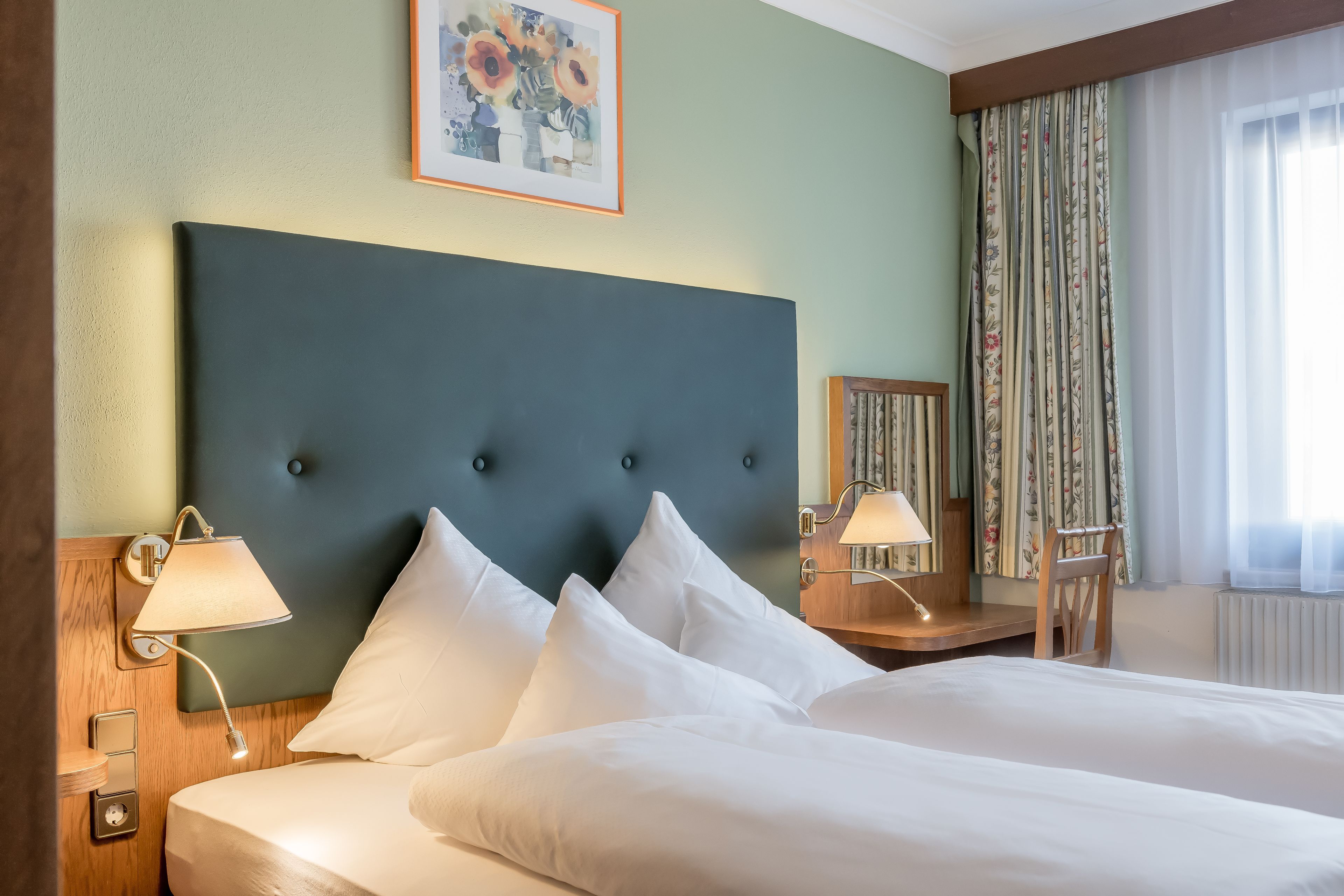 A hotel room with a large bed, two bedside lamps, a headboard, a framed artwork, a wooden nightstand, and a chair.