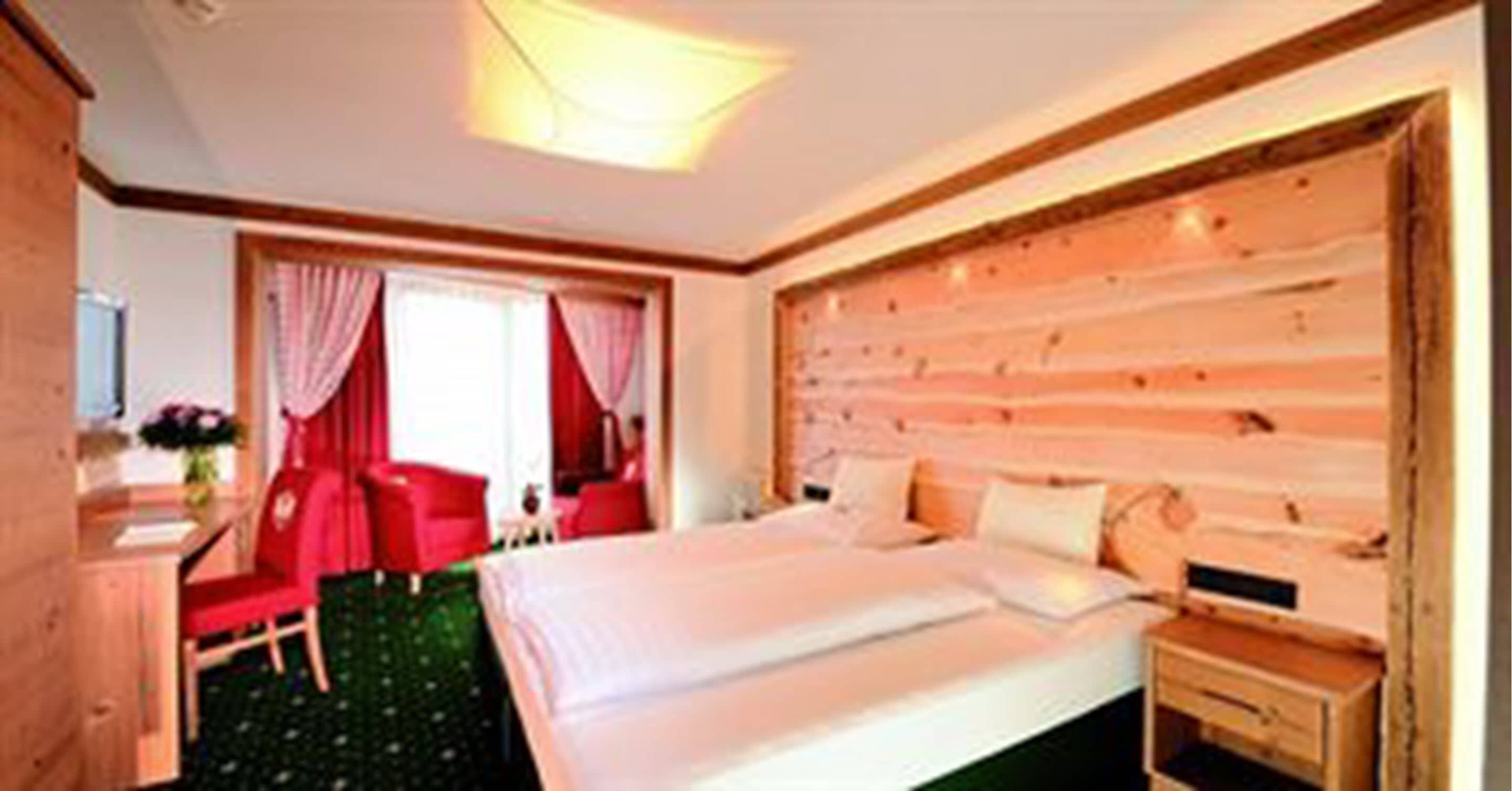 A hotel room with a wooden headboard, a bed with white linens, a desk with a chair, and a window with red curtains.