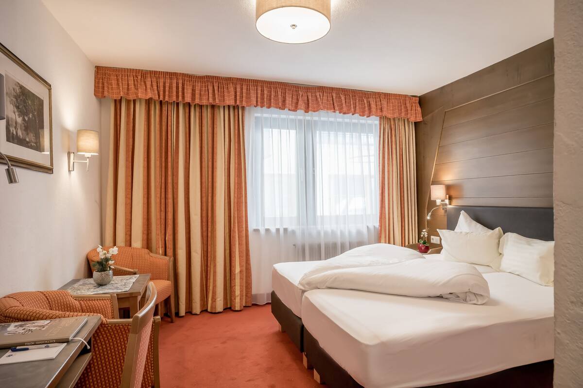 Une chambre d’hôtel avec un lit, un bureau et une chaise, une lampe et une fenêtre avec des rideaux.