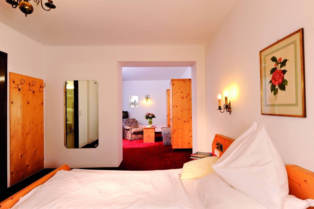 Une chambre d’hôtel avec un lit, une armoire en bois, un miroir, une petite table avec un vase et une porte donnant sur une autre pièce.