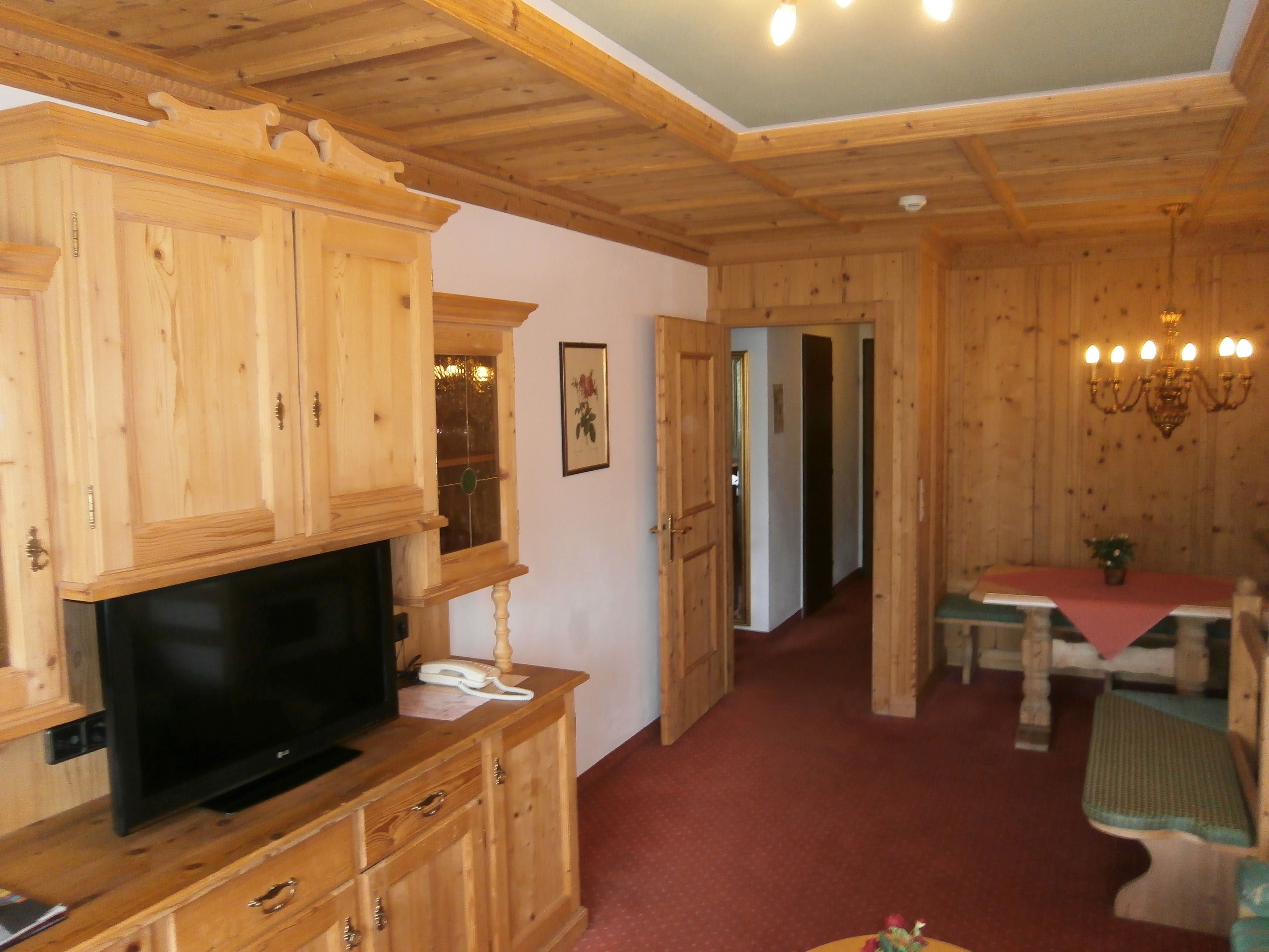 A wooden interior with a TV on a cabinet, a dining table with chairs, and a chandelier.