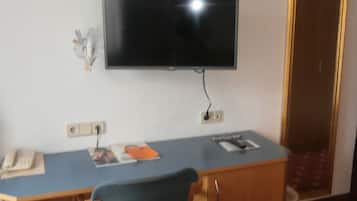 A room with a flat-screen TV mounted on the wall, a desk with a chair, a telephone, and a small table with a folder and a book.