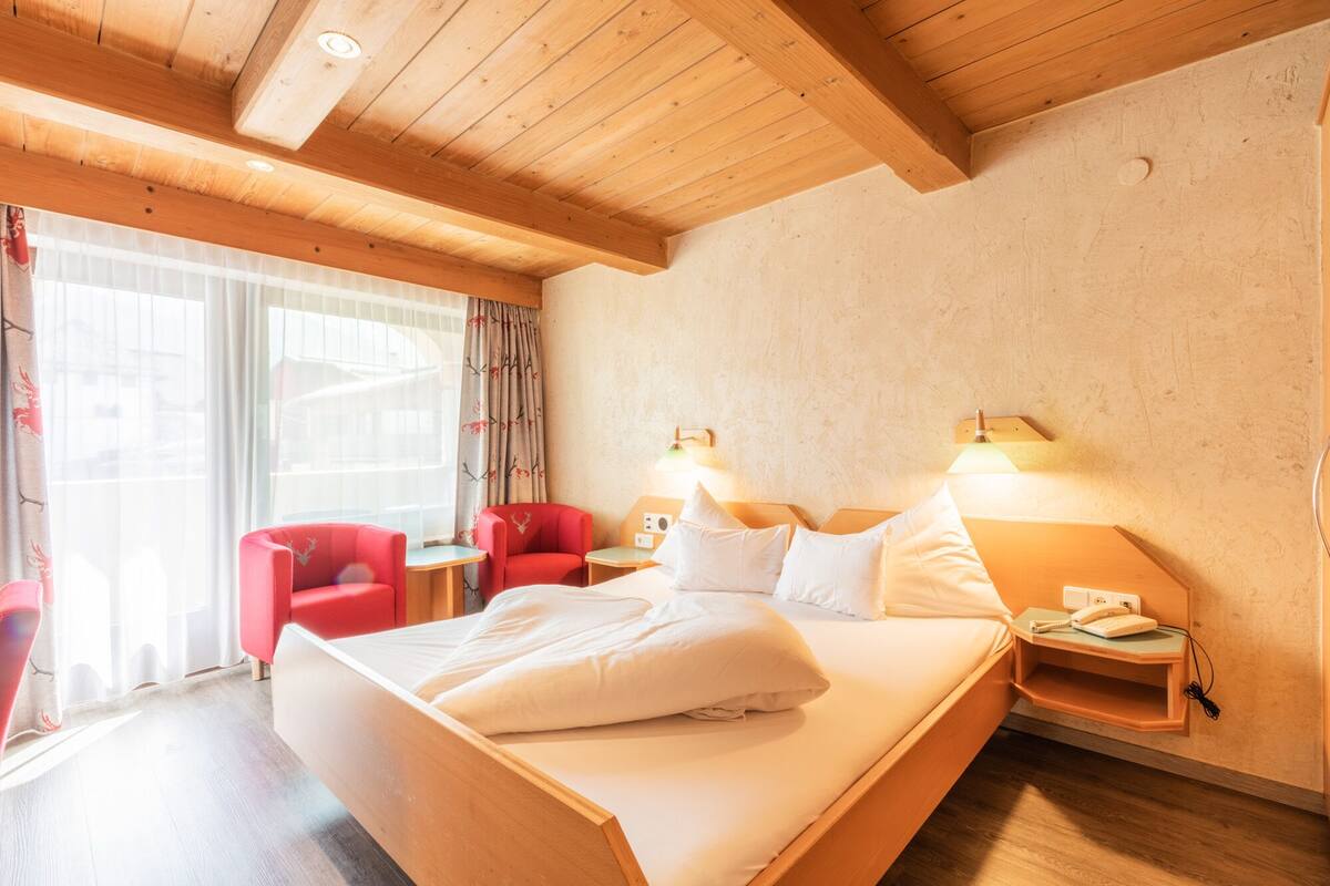 Une chambre d’hôtel avec un plafond en bois, un grand lit avec une literie blanche, deux chaises rouges, une petite table et une lampe.