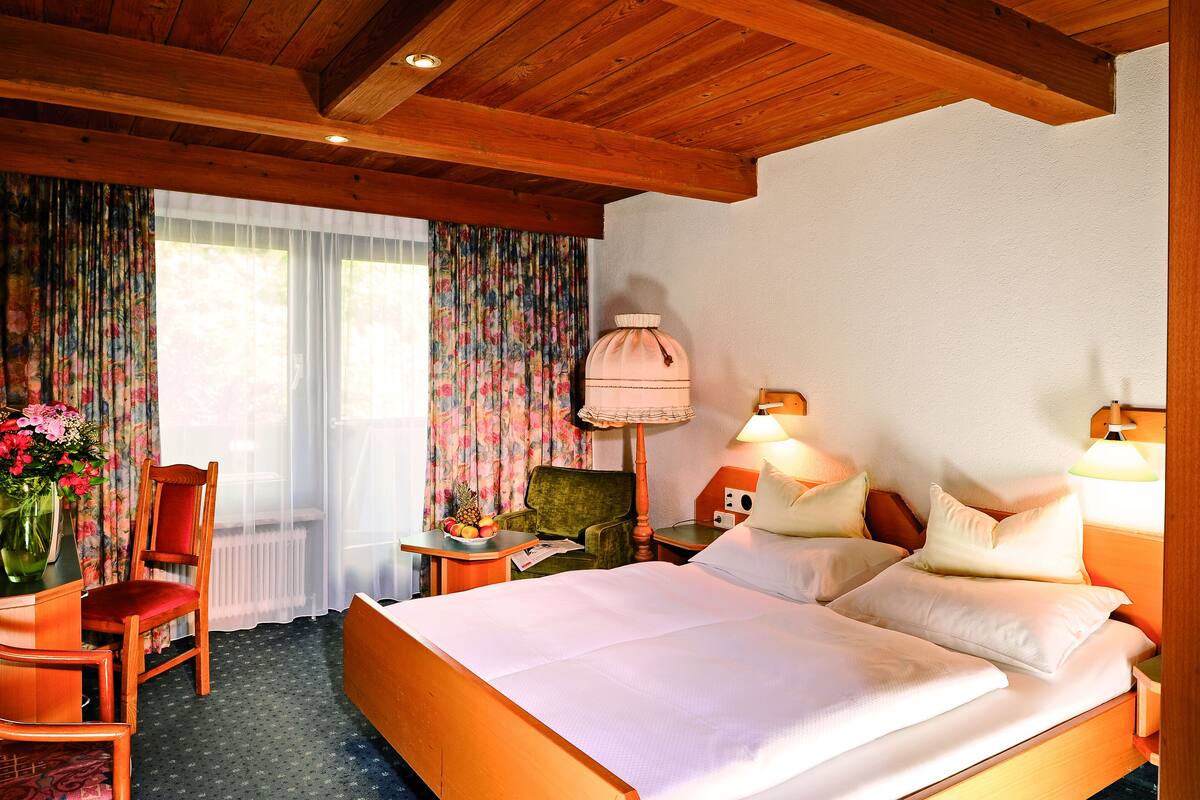 Une chambre d’hôtel avec un plafond en bois, un lit recouvert de draps blancs, un fauteuil vert, une table d’appoint en bois avec une lampe, et un rideau à motifs floraux.