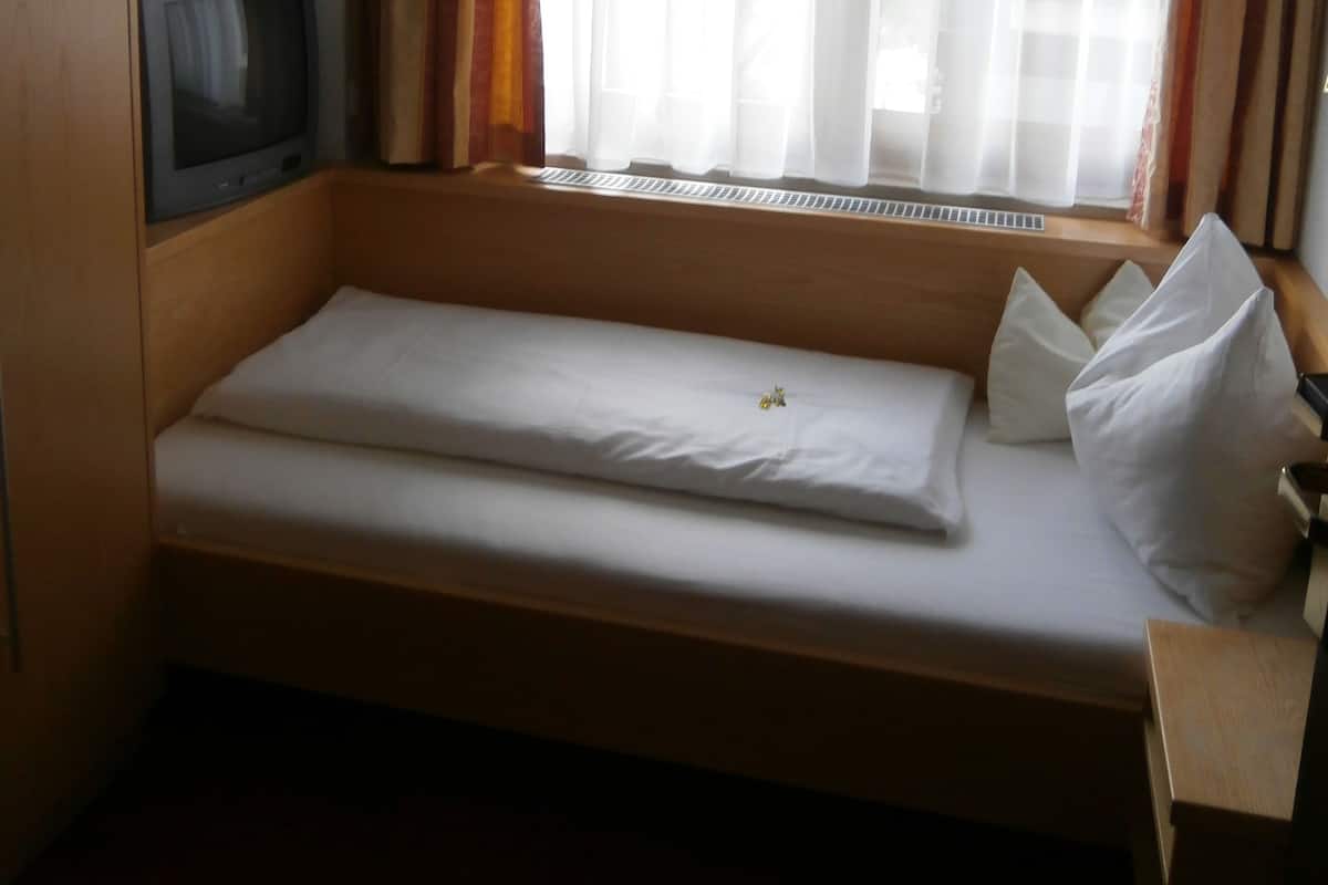 Une petite chambre avec un lit simple, une tête de lit en bois, un téléviseur posé sur un meuble en bois et une fenêtre ornée de rideaux légers.